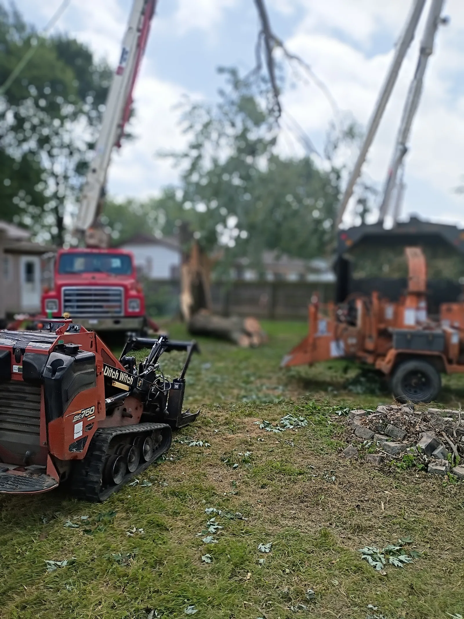 A tracked skid steer loader and a red flatbed truck parked on grass at a job site with a crane boom overhead, an orange wood chipper to the right, and cut logs and debris on the ground.